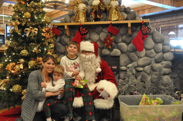 Eine Frau, Kinder und ein Mann in Santa-Klaus-Kostüm mit Brille und Handschuhen stehen neben einem geschmückten Weihnachtsbaum, einer Felswand, Spielzeugen und einem Korb mit Gegenständen, mit Lichtern im Hintergrund.