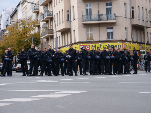 Eine Gruppe von Polizisten in schwarzen Uniformen hält blaue Helme in der Mitte einer Straße, die von Laternenpfählen, Bäumen und Gebäuden mit Glasfenstern und Balkonen gesäumt ist, mit mehr Bäumen und einem klaren blauen Himmel im Hintergrund.