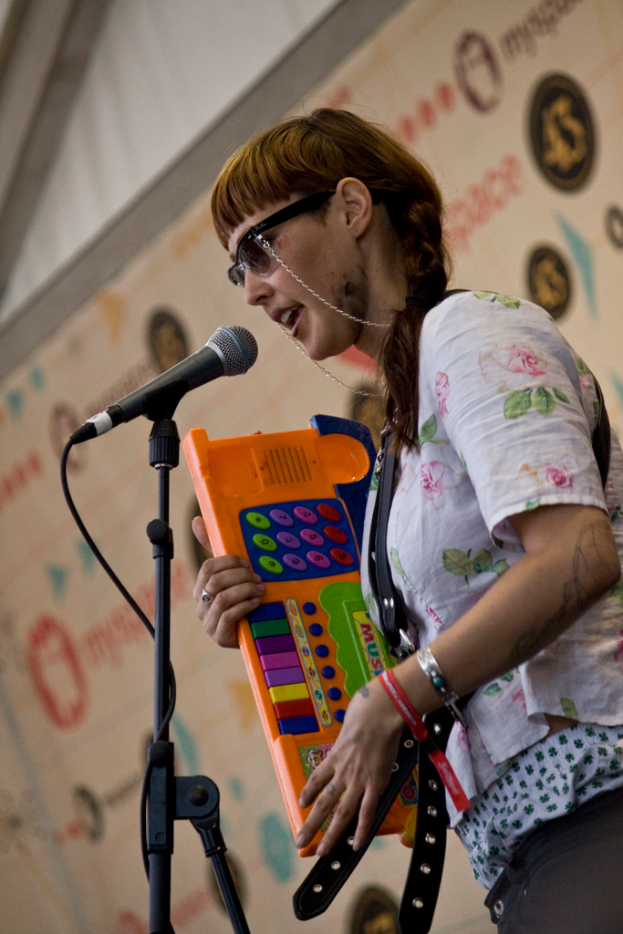 Eine Frau in einem weißen Hemd und Sonnenbrille singt in ein Mikrofon, während sie ein Piano spielt, mit einem Plakat im Hintergrund.