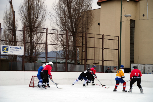 Menschen beim Eishockey auf einem Eis mit Gebäuden, Bäumen, einer Straßenlaterne, einem Namensschild und Zäunen im Hintergrund unter dem Himmel.