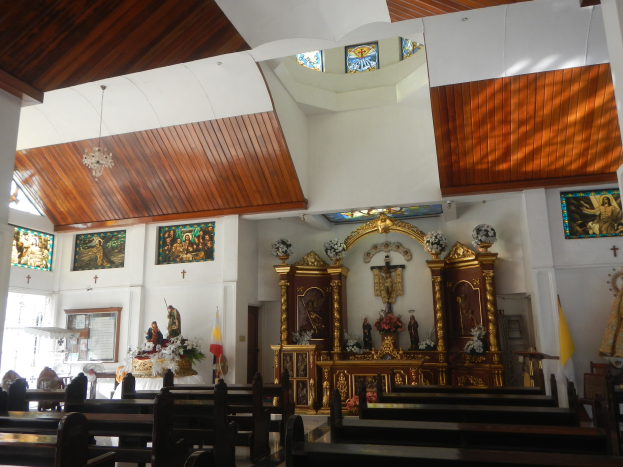 Innere einer Kirche mit hölzernen Kirchenbänken, Statuen, Blumenbouquets, Fahnen, Fotorahmen an den Wänden und Buntglasfenstern, mit Deckenleuchten, die eine gemütliche Atmosphäre schaffen.