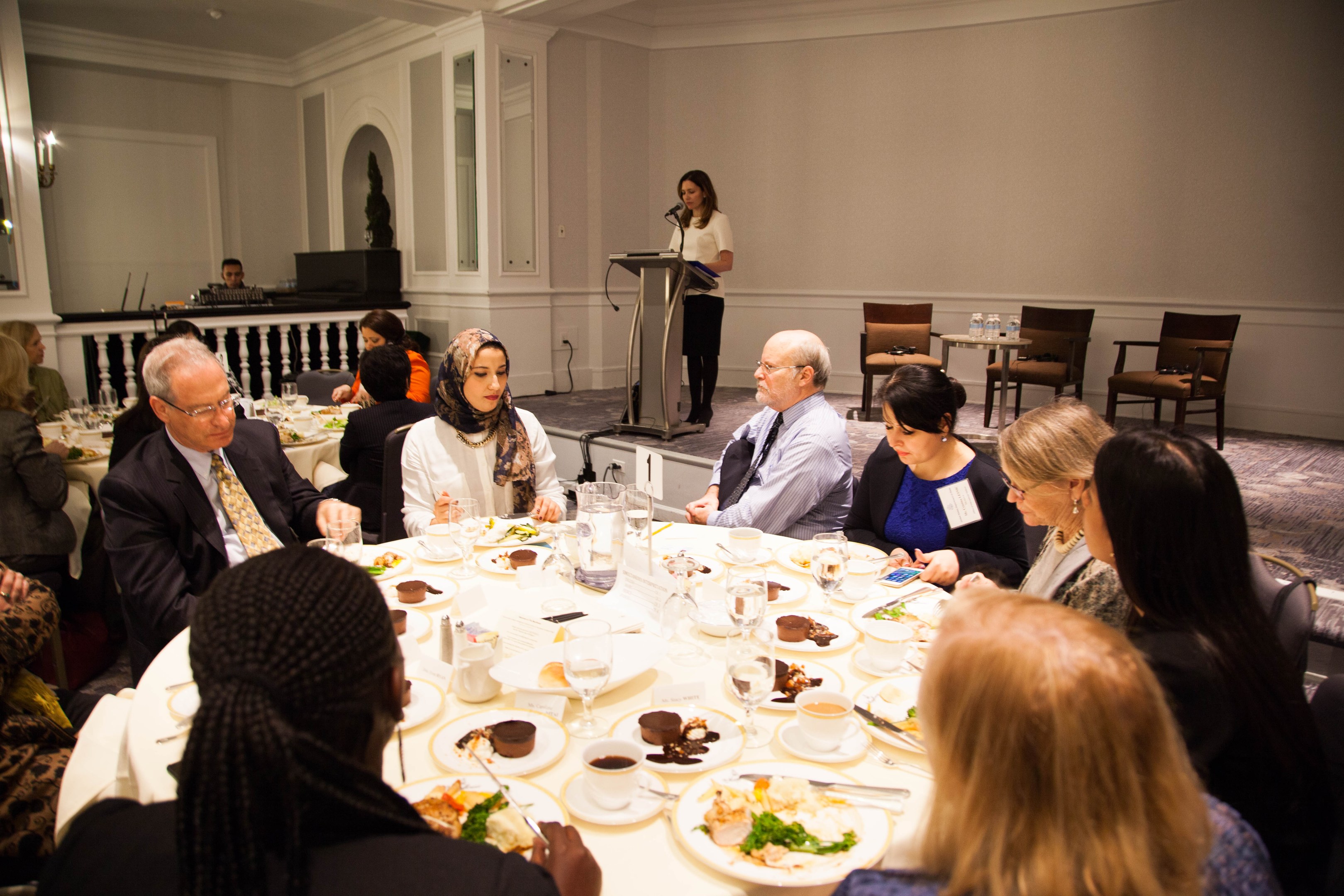 Eine Gruppe von Menschen sitzt um einen Esstisch mit Essen und Besteck, während eine Frau in der Nähe eines Podiums mit einem Mikrofon im Hintergrund steht.