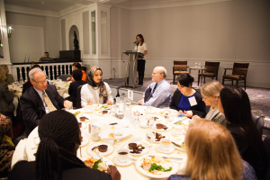 Eine Gruppe von Menschen sitzt um einen Esstisch mit Essen und Besteck, während eine Frau in der Nähe eines Podiums mit einem Mikrofon im Hintergrund steht.