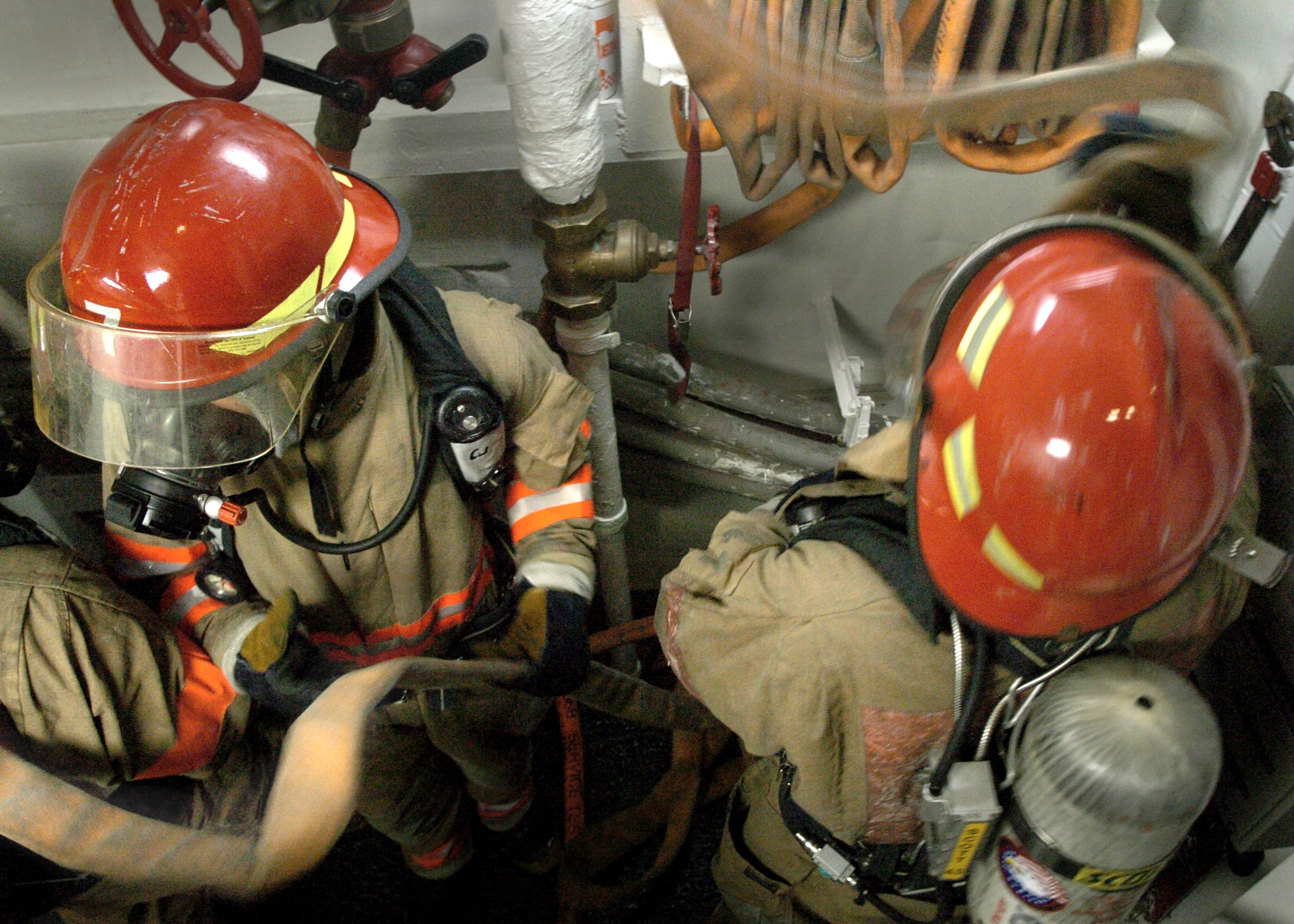 Feuerwehrleute in Schutzausrüstung bei der Arbeit an einem Feuerhydranten, mit Rohren, Ventilen und einer Wand im Hintergrund und Text unten.