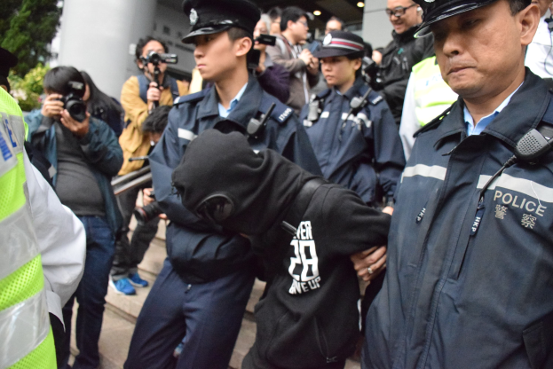 Eine Gruppe von Polizeibeamten steht vor einer Menge von Menschen, einige tragen Mützen und halten Kameras, mit einem Gebäude mit Säulen und Deckenleuchten im Hintergrund und Bäumen links.