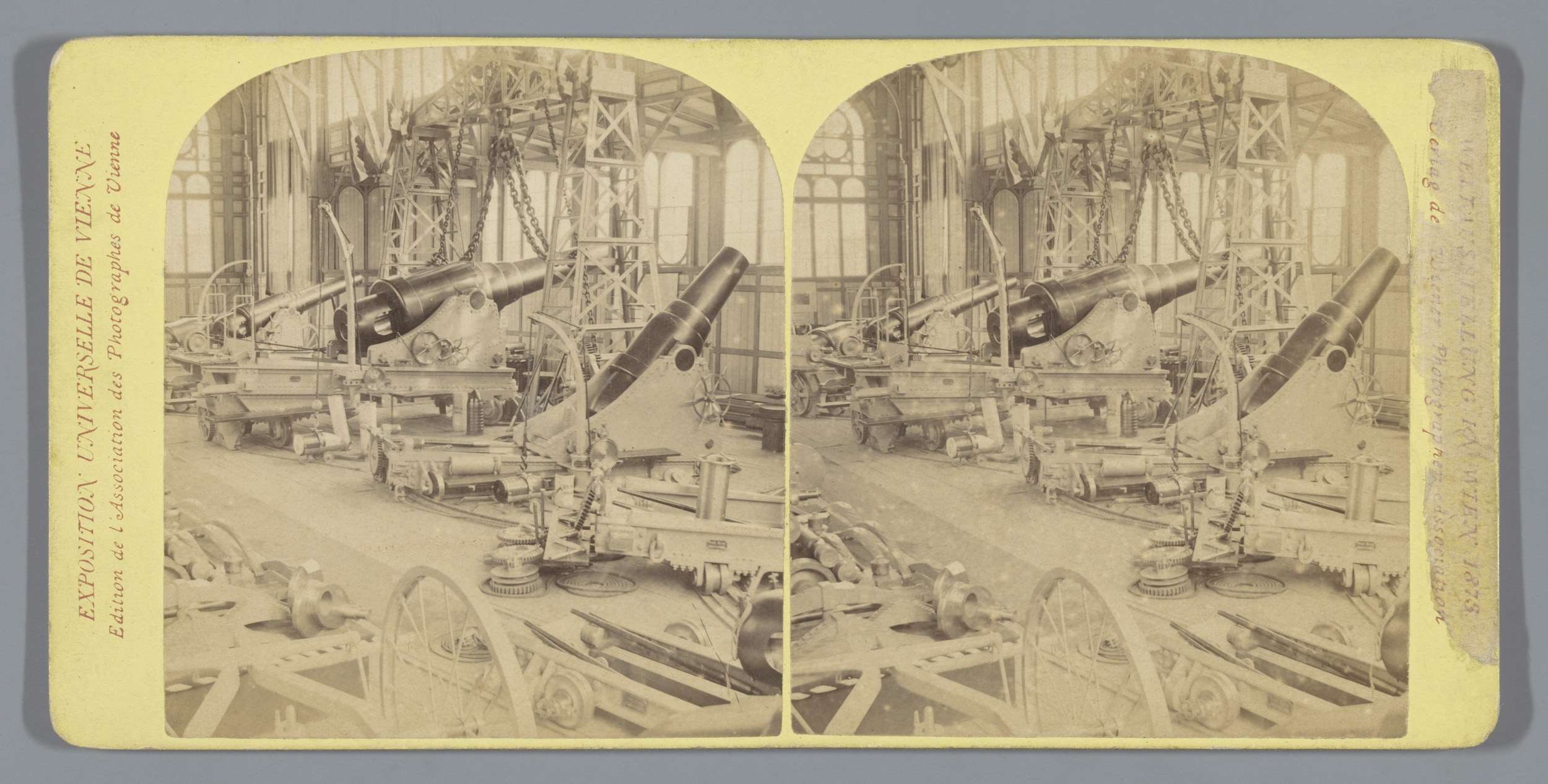 Schwarzes und weißes Foto eines alten Fabrikinneren mit einem gro├čen Geschütz in der Mitte, umgeben von kleineren Geschützen und Metallstangen auf dem Boden, und lesbarem Text auf der linken Seite.