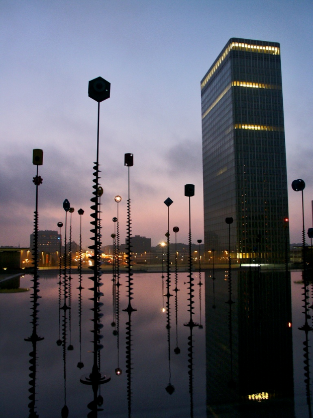 Mehrere Pole und Gebäude mit Lichtern unter einem bewölkten Himmel, mit Spiegelungen der Pole.