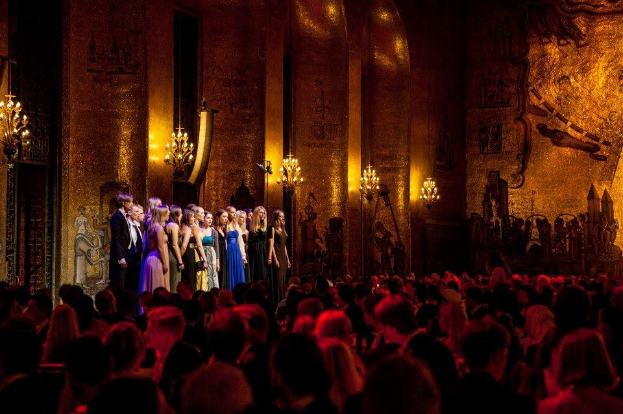 Eine Gruppe von Menschen auf einer beleuchteten Bühne mit bemalten Wänden an der Royal Opera House, betrachtet von einem Publikum.