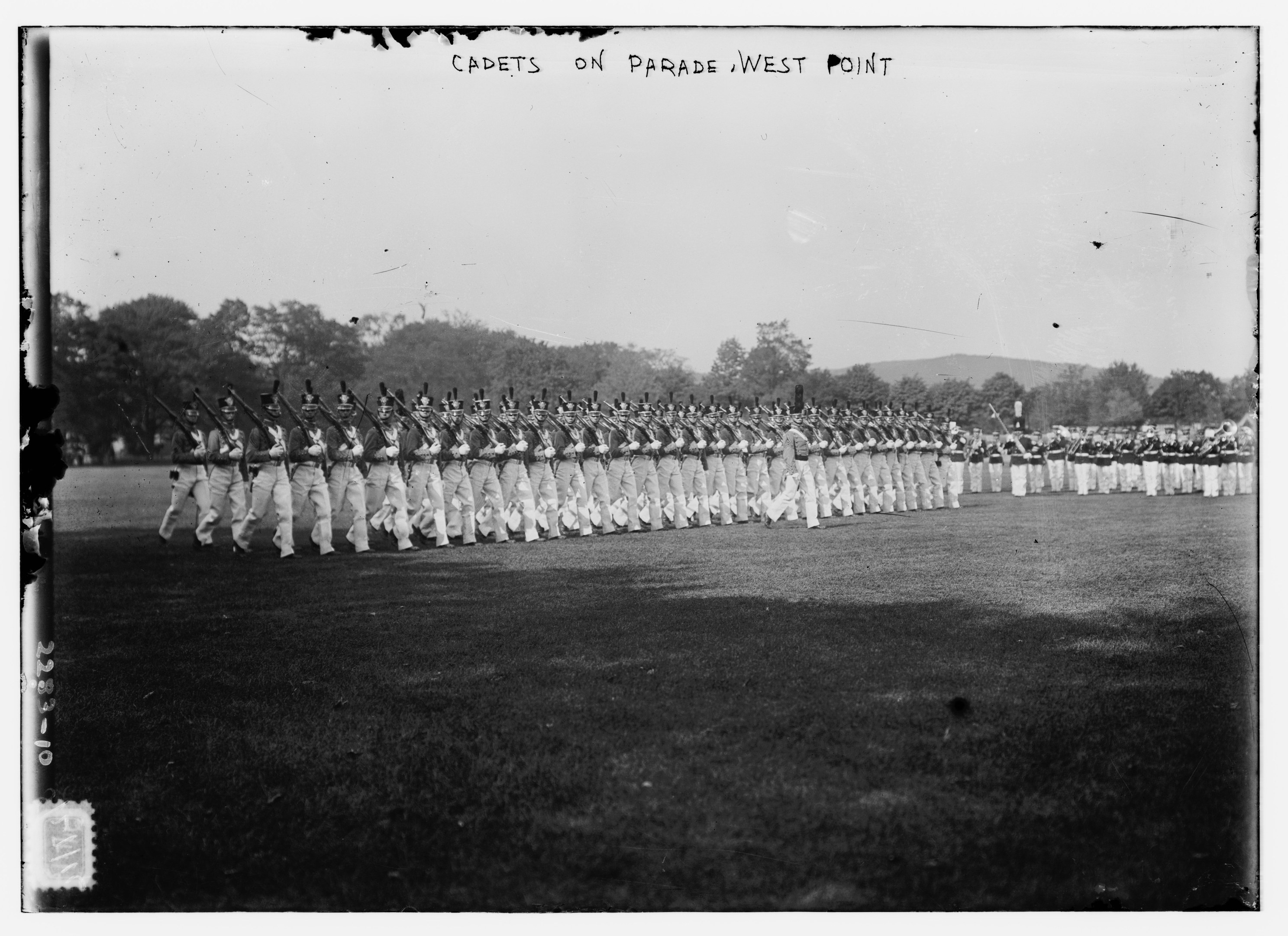 Cadets on parade at West Point, some holding rifles, with trees, hills, and a cloudy sky in the background.