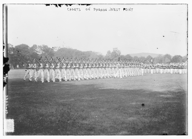 Cadets on parade at West Point, some holding rifles, with trees, hills, and a cloudy sky in the background.