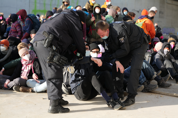 Eine Gruppe von Menschen, einige mit Mützen und Masken, sitzt auf dem Boden mit zwei Polizeibeamten in der Nähe in einer Stadtumgebung, was darauf hindeutet, dass sie festgenommen werden.