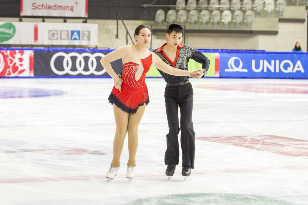 Zwei Eiskunstläufer, ein Mann und eine Frau, gleiten Hand in Hand über eine Eisfläche, mit Zuschauern und Event-Elementen im Hintergrund.