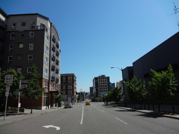 Eine Stadtansicht mit Gebäuden, einer Straße mit Fahrzeugen, Bäumen und Pfosten entlang des Gehwegs unter einem Himmel.