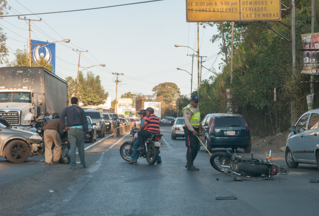 Gruppe von Menschen um ein verunglücktes Motorrad auf dem Seitenstreifen mit vorbeifahrenden Autos und Lastwagen, umgeben von Bäumen, Strommasten, Straßenlaternen und Schildern unter einem klaren Himmel.