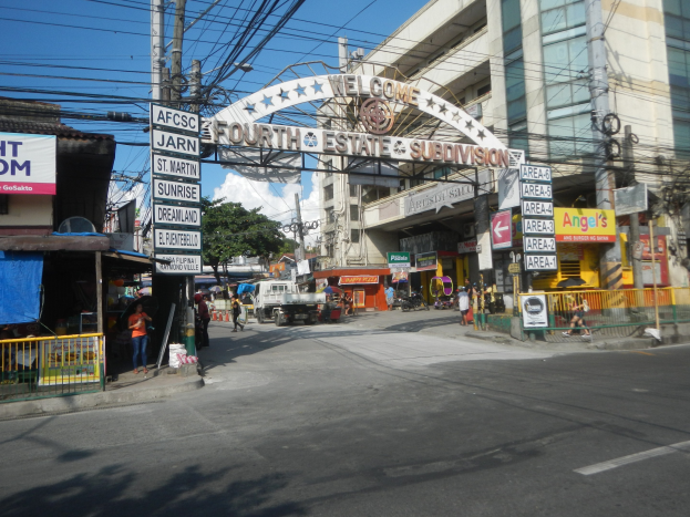 Eine belebte Stadtstraße mit fahrenden Fahrzeugen, Fußgängern auf dem Gehweg, einem Torbogen mit Text, Gebäuden mit Fenstern, Strommasten und -leitungen, Bäumen und einem bewölkten Himmel, mit einem "Fourth Estate Subdivision"-Schilder in der Mitte.