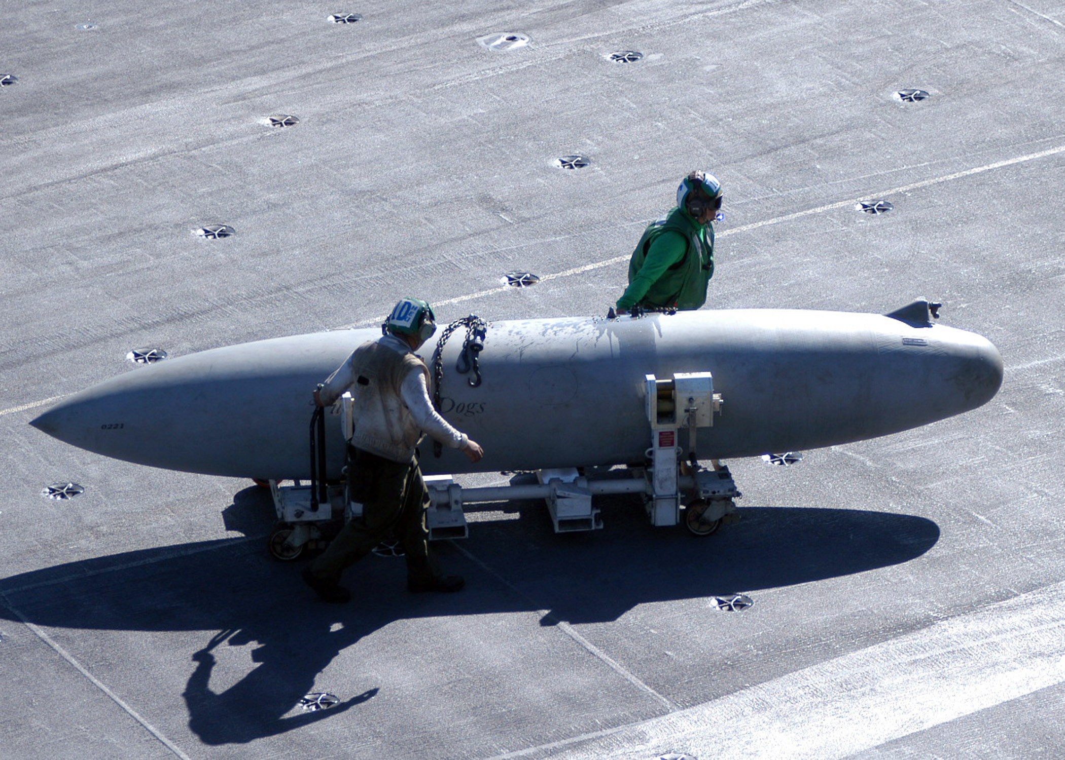 Zwei Männer in Schutzausrüstung inspizieren eine große, spitz zulaufende zylindrische Rakete auf einem Flugzeugträger.