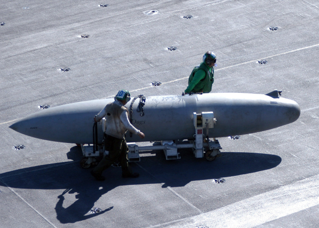 Zwei Männer in Schutzausrüstung inspizieren eine große, spitz zulaufende zylindrische Rakete auf einem Flugzeugträger.