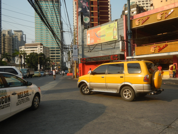 Ein gelbes Taxi fährt auf einer Straße mit hohen Gebäuden, Passanten auf dem Gehweg, Strommasten mit Drähten, Bäumen am Straßenrand und einem sichtbaren Himmel im Hintergrund.