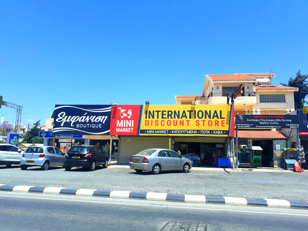 Ein internationales Discounter-Ladenlokal an einer Straßenecke mit verschiedenen Gebäuden, Fahrzeugen, Verkehrszeichen, Straßenlaternen und Bäumen unter einem klaren blauen Himmel.
