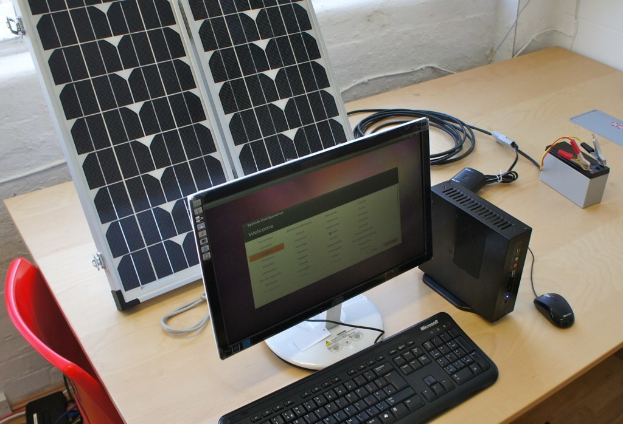 Ein Computersystem mit Tastatur, Maus und CPU auf einem Tisch, neben einem Solarpanel, Akku, Chips und Kabel, mit einem roten Stuhl und einer weißen Wand im Hintergrund.