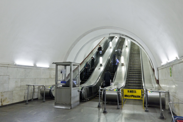 Rolltreppe in einer U-Bahn-Station mit Menschen, Geländern, Informationsschildern, Wandlampen und einem Einkaufswagen am unteren Ende.