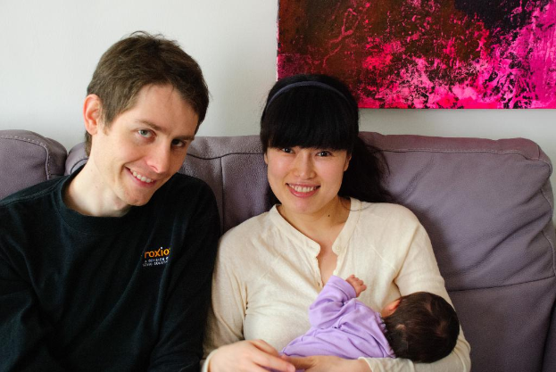 Eine Frau in weißem Hemd hält ein Baby und lächelt, waßhrend sie auf einem Sofa sitzt neben einem Mann in schwarzem T-Shirt, der ebenfalls lächelt, mit einem pinken Fotoраhmen an der weißen Wand dahinter.