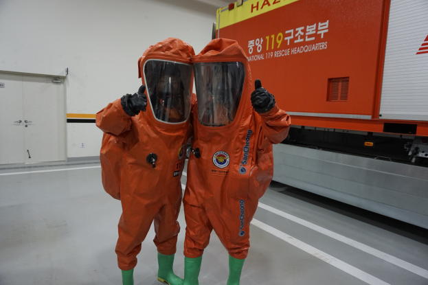 Zwei Personen in orangefarbenen Schutzanzügen stehen nebeneinander vor einem Truck, mit einer Wand und Türen im Hintergrund.
