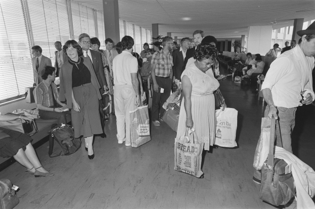 Gruppe von Menschen in einer schwarz-weißen Flughafen-Szene, einige sitzen auf Bänken und andere stehen mit Gepäck, Fenster mit Jalousien links; ein Mann und eine Frau in der Mitte halten Taschen nebeneinander.
