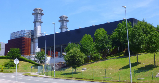 Großes Gebäude mit zahlreichen Fenstern, als Kraftwerk identifiziert durch Beschilderung, umgeben von Straßeninfrastruktur, Fahrzeugen, Grünflächen und einem bewölkten Himmel.