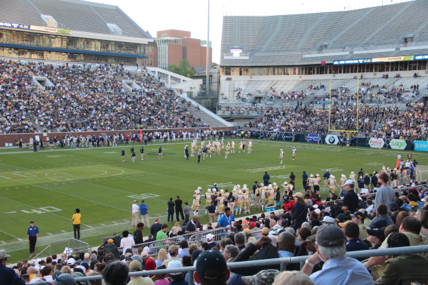 Fußballspiel in einem Stadion mit Spielern auf dem Feld und Zuschauern in den Rängen unter einem klaren blauen Himmel.