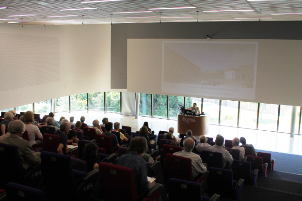 Ein Seminarsaal in einer Universität mit vielen Menschen, die aufmerksam einer Präsentation an der Front zugewandt sind, wo eine Person an einem großen Tisch die Präsentation erklärt, mit Glasfenstern im Hintergrund, die Bäume und Gebäude zeigen.