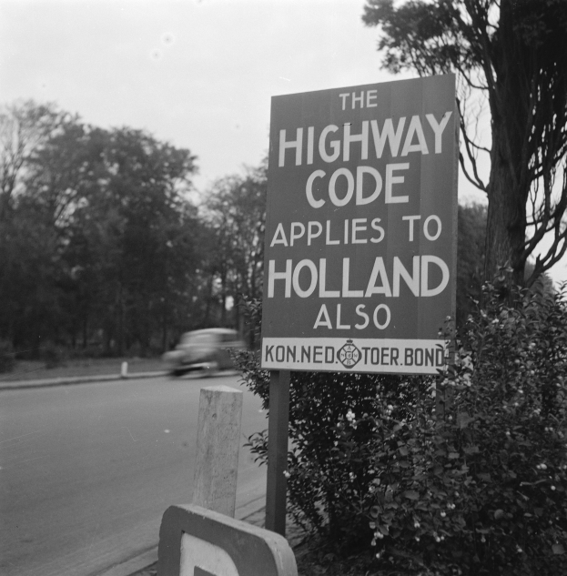 Ein Schwarz-Weiß-Foto eines Straßenschilds mit der Aufschrift "Die Straßenverkehrsordnung gilt auch für Holland" mit einem vorbeifahrenden Fahrzeug, umgeben von Bäumen und einem klaren Himmel.