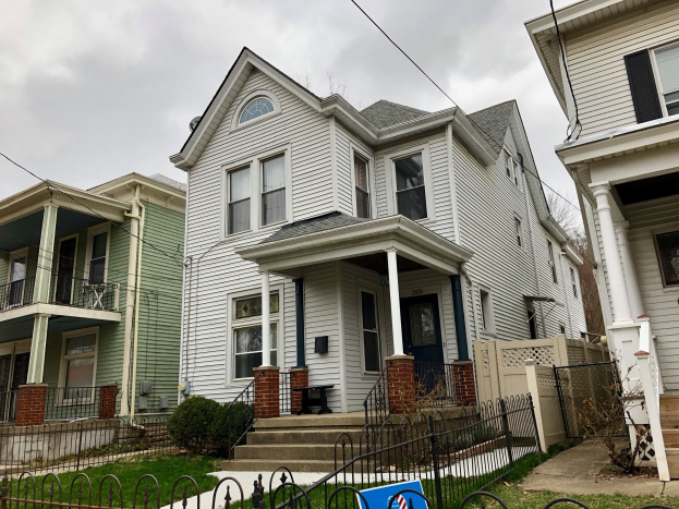 Reihe von Häusern mit Fenstern, Türen, Säulen, Geländern, Treppen und Dächern, umgeben von einem Metallzaun, Gras, Pflanzen, Bäumen und einem bewölkten Himmel, mit einem Mietschild am Boden, das die Adresse und die Listen Details anzeigt.