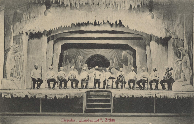 Ein Schwarz-Weiß-Foto einer Gruppe von Menschen auf einer Bühne mit Musikinstrumenten, Treppen, die zur Bühne führen, und Skulpturen auf beiden Seiten, mit dem Text "Lindenhof, Zittau, Deutschland" unten.