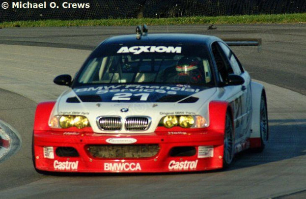 Eine Person mit Helm fährt einen BMW M3 GT3 Rennwagen auf einer von Gras und einem Maschendrahtzaun umgebenen Rennstrecke.