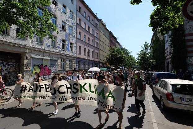 Eine Gruppe von Menschen marschiert bei einer Demonstration, hält ein Banner, mit parkenden Autos auf der rechten Seite, einem Radfahrer auf der linken Seite, Gebäuden, Bäumen und einem Schild im Hintergrund unter einem sichtbaren Himmel.