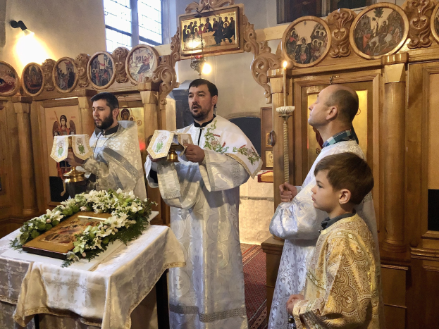 Eine Gruppe von Menschen um einen Tisch in einer Kirche herumstehend, Bücher haltend, mit einem Tuch und einem Blumenstraß auf dem Tisch und gerahmten Fotos, Lichtern, einem Fenster und einem hölzernen Gegenstand im Hintergrund.