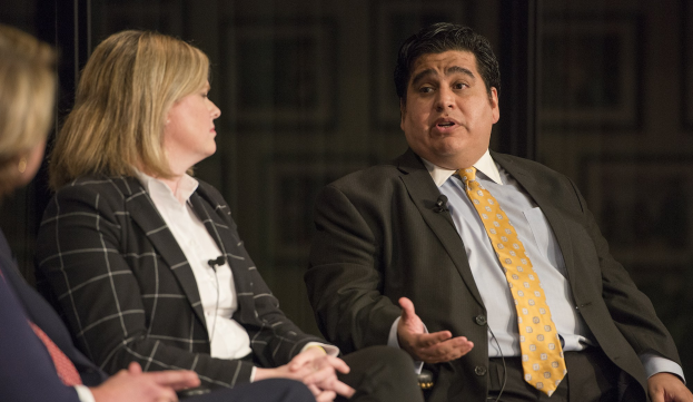 Zwei Personen in Anzügen sitzen nebeneinander auf einer dunklen Bühne, der Mann trägt ein schwarzes Sakko mit weißem Hemd und gelber Krawatte.