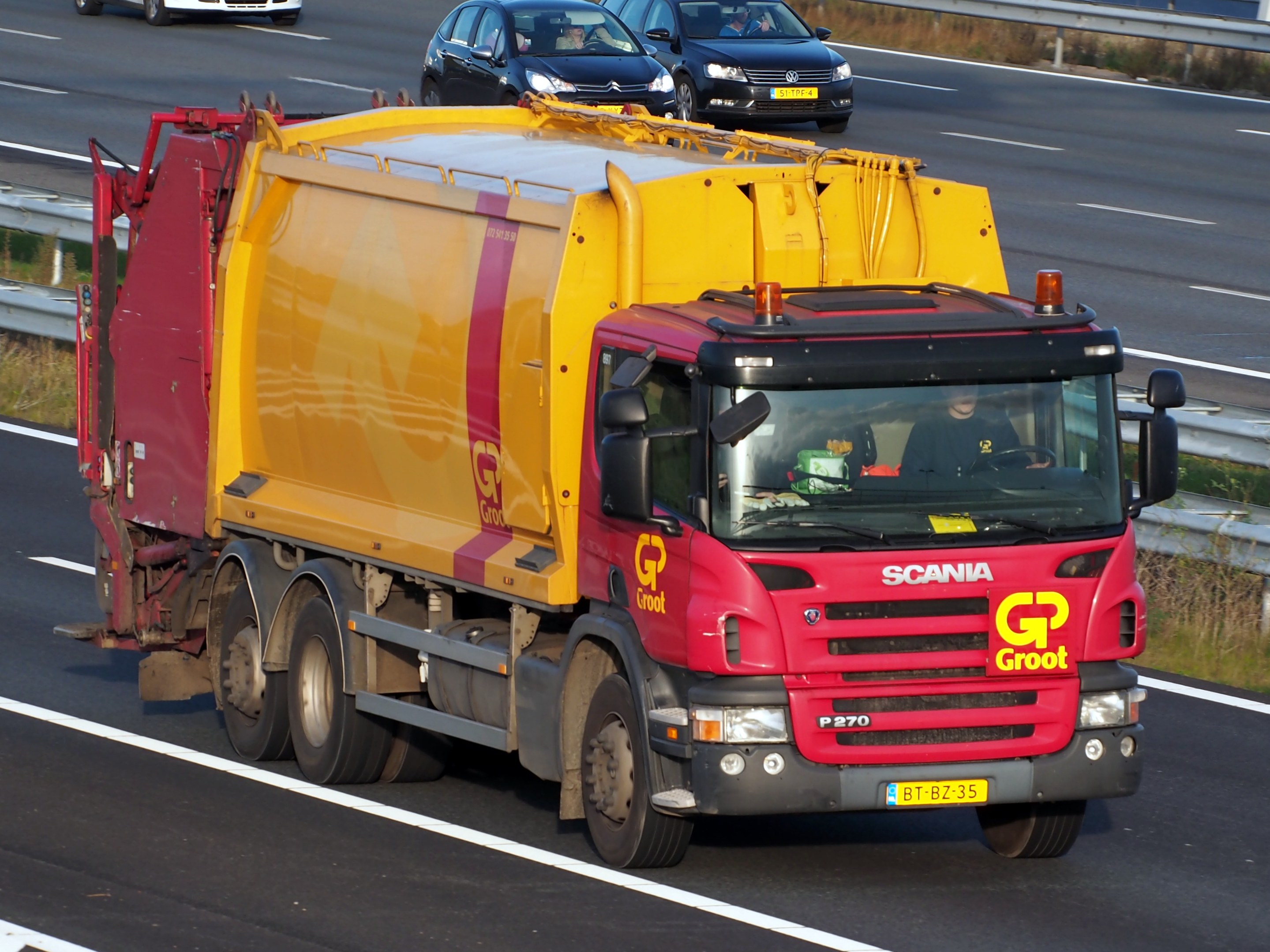 Ein gelber und roter Müllwagen auf einer Autobahn umgeben von Autos, mit Gras und einer Leitplanke auf beiden Seiten der Straße.