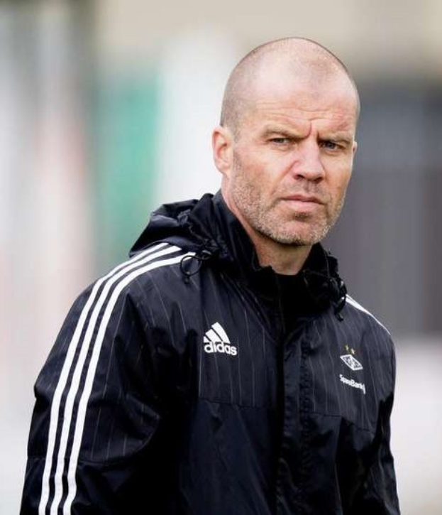 Kahlköpfiger Trainer in einer blauen Jacke mit weißen Streifen und weißem Kragen, der auf einem Fußballfeld steht und nachdenklich dreinschaut.