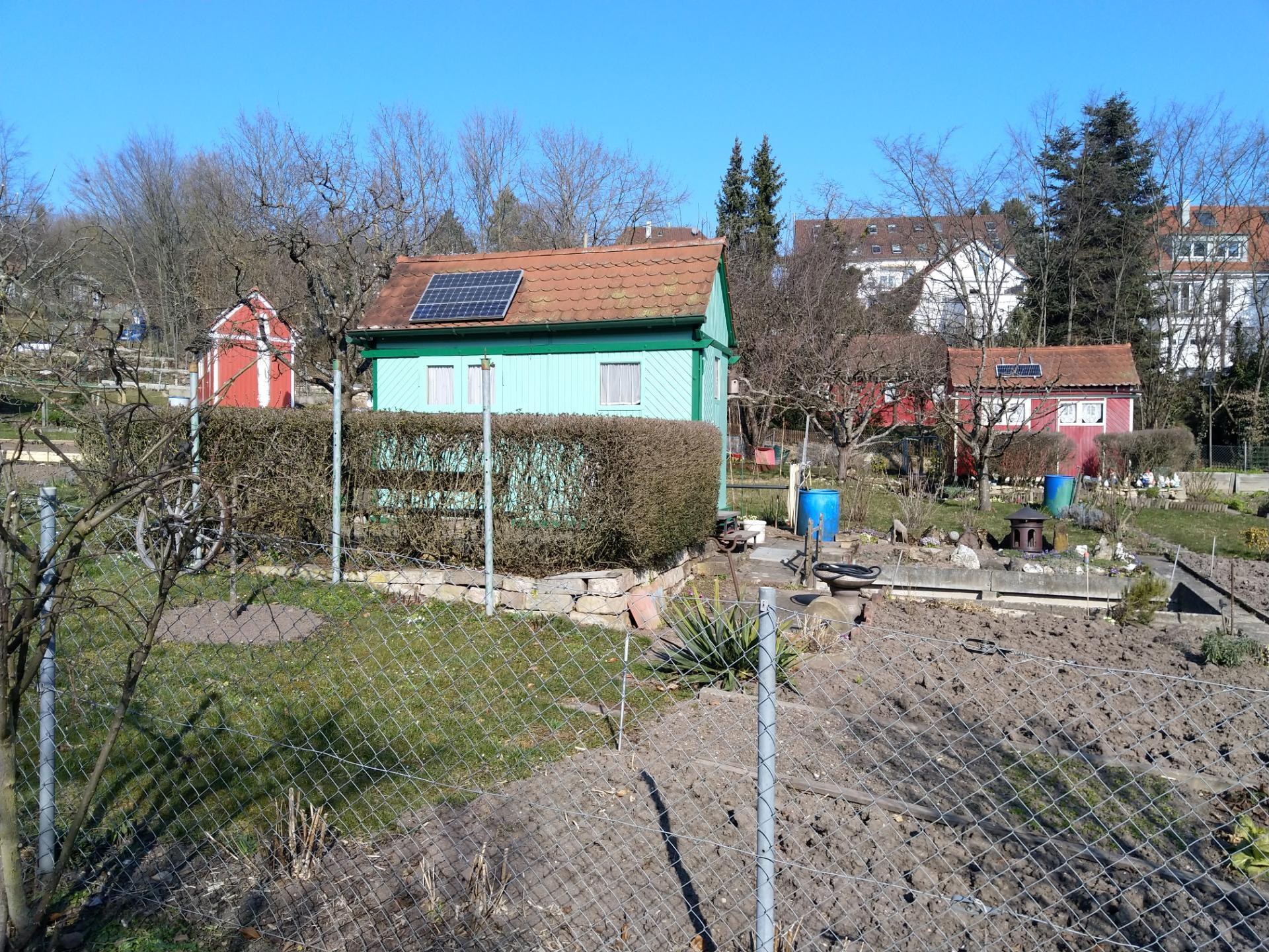Kleines grünes Haus mit Solarpanelen auf dem Dach, umgeben von Maschendraht, vor einem klaren blauen Himmel mit Bäumen, Pflanzen und Gras im Vordergrund.