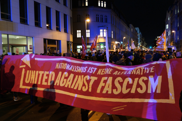 Eine Gruppe von Menschen marschiert mit einem Banner, auf dem 'Einheit gegen Faschismus' steht, eine Straße entlang, mit Gebäuden, Laternenmasten und Schildern am Straßenrand und dem Himmel im Hintergrund.