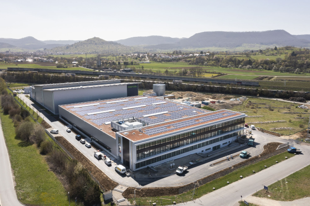 Luftaufnahme eines großen Gebäudes mit Solarmodulen auf dem Dach, umgeben von Fahrzeugen, Bäumen, Gras und Masten, mit Hügeln und einem klaren blauen Himmel im Hintergrund; das Gebäude ist Deutschlands erste solarbetriebene Fabrik.
