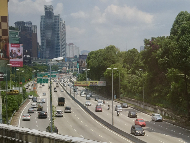 Vielbefahrene Autobahn mit hohen Gebäuden, Straßeninfrastruktur, Bäumen und bewölktem Himmel im Hintergrund.
