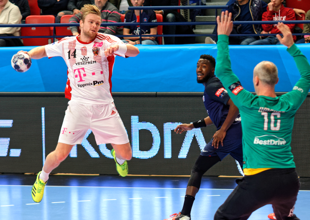 Eine Gruppe von Männern, die Handball auf einem Platz spielen, mit einem Ball in der Mitte der Aktion, während Zuschauer von Stühlen und Geländern in einer Arena zuschauen. Das Anzeigebrett im Hintergrund zeigt "Futsal-Weltmeisterschaft 2019 - Bayern München gegen Paris Saint-Germain".