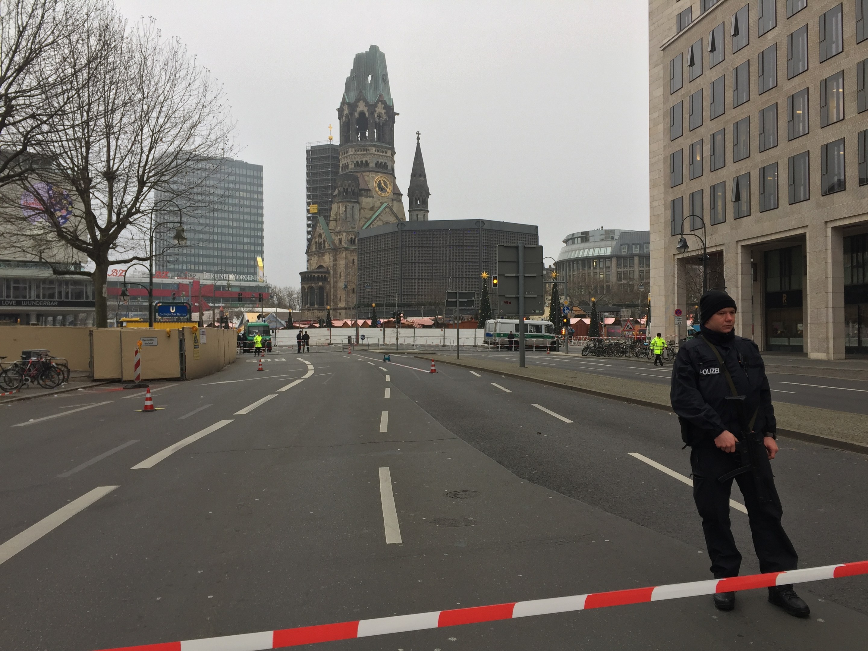 Ein Polizist in schwarzer Uniform und Mütze steht auf einer Straße in Berlin umgeben von Absperrbaken, Fahrrädern, Laternen, Bäumen und Gebäuden, im Hintergrund ist ein Kirchturm und ein klarer blauer Himmel zu sehen.