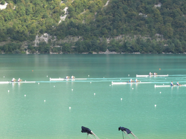 Eine Gruppe von Menschen bei einem Ruderwettbewerb auf einem Gewässer mit Bäumen und einem Hügel im Hintergrund, sowie Kleidung über Tribünen am Boden.
