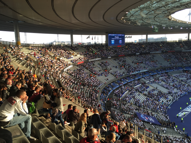 Eine gro\u00dfe Menge von Menschen sitzt im Allianz Stadion in M"unchen, Deutschland, bei einem Fuballspiel, mit einer B"hne auf der rechten Seite und Fahnen, Masten und einem Bildschirm im Hintergrund.