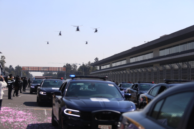 Eine Gruppe von Polizeiwagen vor einem Gebäude geparkt, mit Menschen auf der linken Seite der Straße und vier Hubschraubern am Himmel.