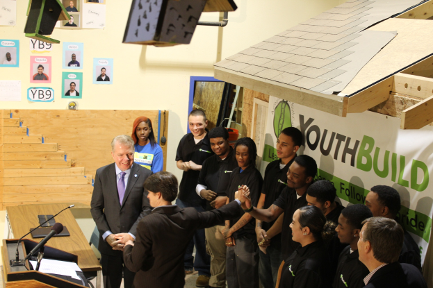 Eine Gruppe von Menschen, die um ein Rednerpult herum stehen und sich die Hände schütteln, vor einem Publikum, mit Mikrofonen und Papieren auf einem nahen Tisch, vor einer Wand mit Plakaten und Gegenständen auf dem Dach.
